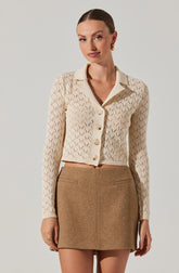 A woman wearing a cream-colored, long-sleeved, button-up cardigan with a diamond-shaped open-knit pattern. The cardigan has a collar and is cropped, ending just above her waist. She pairs it with a high-waisted, brown, woolen mini skirt that has a simple, straight cut and two welt pockets at the front.